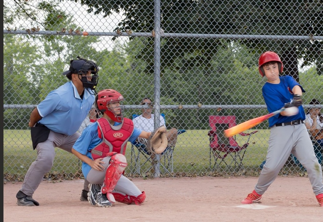 Young baseball player mid-swing in a live game, catcher and umpire behind